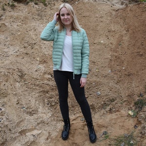 May include: A light green quilted bomber jacket with a front zipper, worn over a plain white t-shirt, paired with dark gray leggings and black lace-up combat boots. The jacket features ribbed cuffs and a high collar, providing a layered look.
