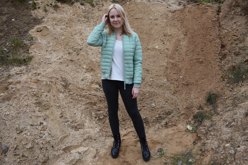 May include: A light green quilted bomber jacket with a front zipper, worn over a plain white t-shirt, paired with dark gray leggings and black lace-up combat boots. The jacket features ribbed cuffs and a high collar, providing a layered look.
