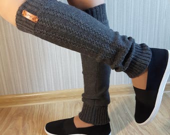 May include: A pair of dark gray knit leg warmers featuring a ribbed texture at the top and bottom cuffs, a central cable knit pattern, and a small rectangular brown leather tag with text attached to the upper portion of the left leg warmer.