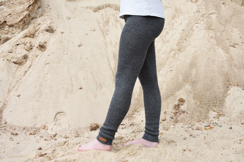 May include: Charcoal gray leggings featuring a fitted silhouette and a small rectangular brown leather-like patch detail at the ankle. The leggings are worn with a plain white t-shirt, extending down to the ankles with a textured knit appearance.