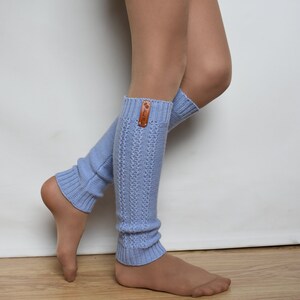 May include: A pair of light blue knit leg warmers featuring a vertical cable-knit pattern and ribbed cuffs at the top and bottom. A small, rectangular brown leather tag with a visible logo is attached to the upper edge of the left leg warmer.