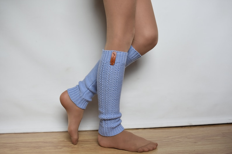 May include: A pair of light blue knit leg warmers featuring a vertical cable-knit pattern and ribbed cuffs at the top and bottom. A small, rectangular brown leather tag with a visible logo is attached to the upper edge of the left leg warmer.