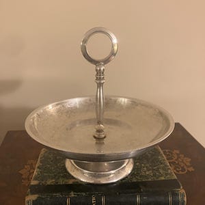 Op de afbeelding: Een zilverkleurige metalen serveerschaal met een ronde kom en een centrale verticale handgreep met een ring aan de bovenkant. De schaal heeft een rand met kraalmotief en rust op een voetstuk. Het metalen oppervlak vertoont slijtage.