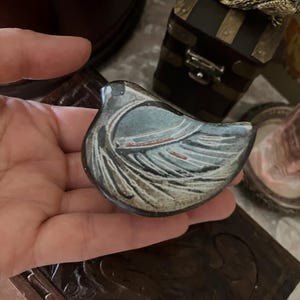 以下が含まれることがあります： 滑らかで丸みを帯びたフォルムの小さな陶器製の鳥の置物。スレートブルー、クリーム色、チャコールグレーの落ち着いた色合いで、表面には羽のような模様が刻まれ、錆びたような色のアクセントが施されています。