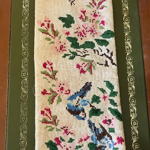 May include: A rectangular piece of cream-colored fabric featuring a cross-stitch embroidery pattern. The design displays three blue birds perched among clusters of pink flowers and green leaves, arranged vertically along the center of the textile.