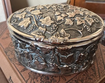 May include: An oval silver-toned metal trinket box featuring a raised relief design of grapevines, leaves, and clusters of grapes. The surface has a textured, cross-hatched background pattern behind the metallic botanical motifs on the lid and sides.
