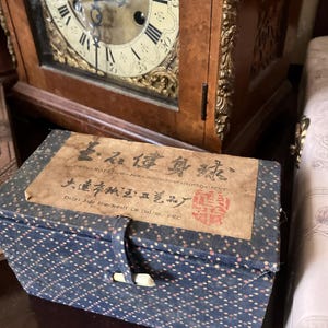 Op de afbeelding: Een rechthoekige blauwe stoffen doos met een kleine witte knevelsluiting en een herhalend patroon van kleine oranje en witte stippen. Een bruin papieren label op het deksel bevat zwarte Chinese karakters, de tekst "TWIN-ROTATING-Jade-BallforHealthIMPROVEMENT" en "Dalyi Jade Handicraft Co Dalian, PRC".
