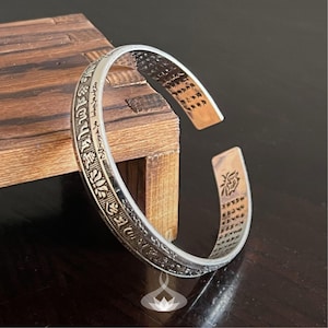 May include: A silver-toned open cuff bracelet featuring intricate embossed script and lotus flower motifs along the outer band. The interior surface displays engraved text and a small lotus flower symbol at one end, with a polished finish.