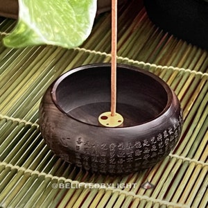 May include: A dark brown wooden incense holder with carved text around the bowl's exterior. A gold-colored metal disc holds an incense stick. The holder sits on a bamboo mat. The text "@BELIFTEDBYLIGHT" is at the bottom.