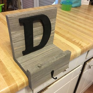 May include: A wooden shelf with a black letter D attached to the front. The shelf is mounted to a wall and has a metal hook underneath. The shelf is made of reclaimed wood and has a rustic look.