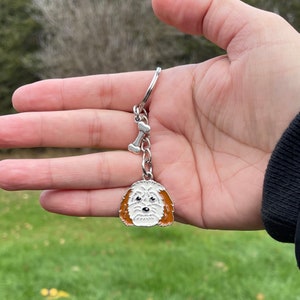 May include: A silver keychain featuring a white dog with brown ears and a silver bone charm. The keychain is held in a hand.