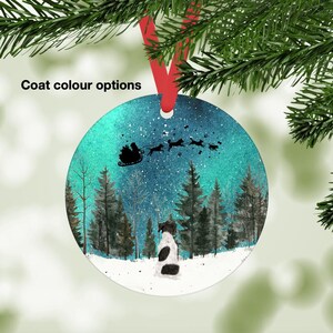 May include: Round Christmas ornament with a winter scene. A dog with black and white markings sits in the snow, gazing at Santa's sleigh pulled by dogs. The background features a forest and a starry sky. The ornament hangs from a red ribbon.