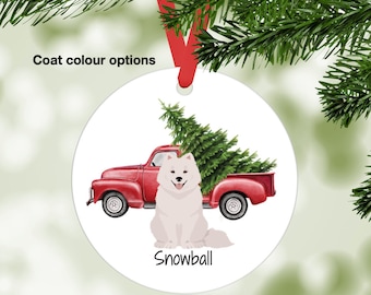 May include: A round, white Christmas ornament featuring a red pickup truck carrying a green Christmas tree. A fluffy white dog named "Snowball" sits in front of the truck. The ornament hangs from a red ribbon. Text on the ornament reads "Coat colour options".