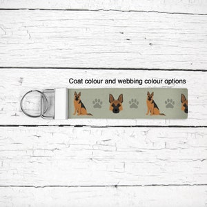 May include: A green and white key chain featuring a German Shepherd dog design. The dog is brown and black with a white chest. The key chain has a silver key ring and a silver clasp. The text "Coat colour and webbing colour options" is printed on the key chain.