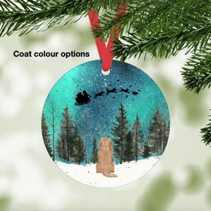 May include: Round Christmas ornament with a winter scene. A light brown dog is looking up at Santa's sleigh pulled by reindeer. The background features a snowy forest under a starry sky. The ornament has a red ribbon for hanging.