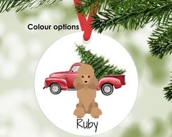 Puede incluir: Un adorno navideño redondo y blanco con una cinta roja. Presenta una camioneta roja que transporta un árbol de Navidad y un caniche marrón llamado "Ruby". El texto "Colour options" está encima del adorno.