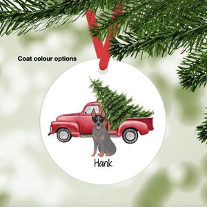 May include: A round Christmas ornament featuring a red pickup truck carrying a Christmas tree. A gray and tan dog named Hank is sitting in front of the truck. The ornament has a red ribbon for hanging.