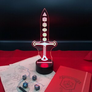 May include: A red and white illuminated sword-shaped lamp with a design of dice on the blade. The lamp is on a black base and is sitting on a table with a map and a red notebook.