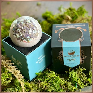 May include: A bath bomb with dried flowers sits on a teal box with the words "Relax, Recharge, Roll". A second box, black with teal accents, reads "Bath Bomb of Bardic Bliss". The items are surrounded by green moss.