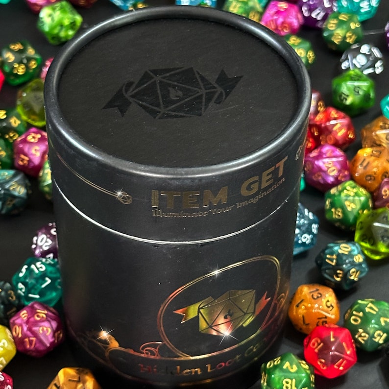 Puede incluir: Un recipiente cil&iacute;ndrico negro con una etiqueta dorada que dice "ITEM GET Illuminate Your Imagination" y una imagen dorada de un dado de 20 caras. El recipiente est&aacute; rodeado de dados de 20 caras de colores.