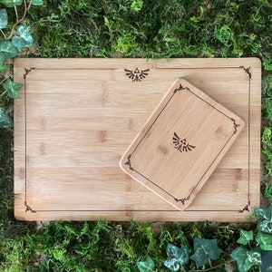 Puede incluir: Una tabla de cortar de bamb&uacute; con una tabla de cortar m&aacute;s peque&ntilde;a insertada. Ambas tablas tienen un borde decorativo y un dise&ntilde;o estilizado en el centro.