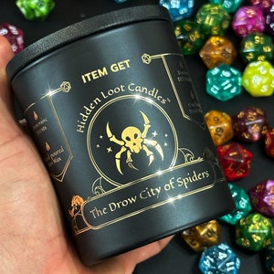 Könnte beinhalten: Schwarzes Kerzengefäß mit goldener Beschriftung, die "ITEM GET Hidden Loot Candles" und "The Drow City of Spiders" lautet. Eine goldene Spinne mit einem Schädel befindet sich in der Mitte des Gefäßes. Das Gefäß ist von bunten Würfeln umgeben.
