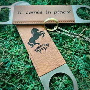 May include: A brown leather bottle opener with a metal ring and a silver metal opener. The bottle opener has the text "It comes in pints?" and a black silhouette of a horse with the text "Prancing Pony".