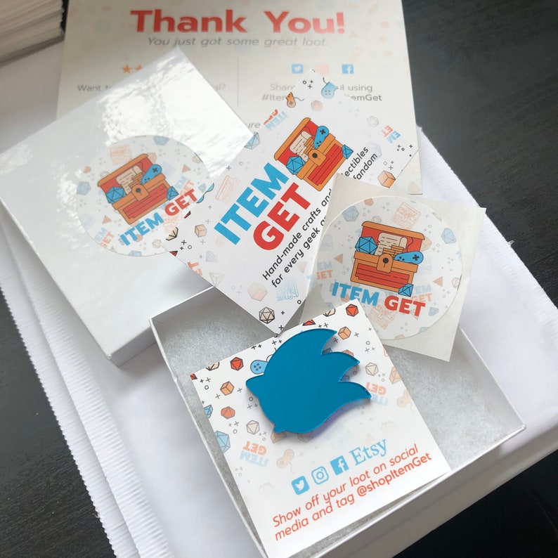 May include: A white box with a blue bird-shaped pin inside, accompanied by stickers and cards. The stickers and cards feature the text "ITEM GET" and a treasure chest graphic. The packaging includes a thank you note.