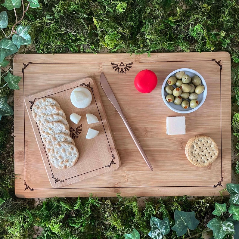 Puede incluir: Una tabla de cortar de madera con un dise&ntilde;o de Triforce, un caramelo rojo, un taz&oacute;n peque&ntilde;o de aceitunas verdes, un cuchillo, galletas saladas y cubos de queso.