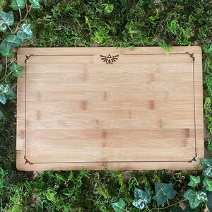 Puede incluir: Una tabla de cortar de bamb&uacute; natural con un borde decorativo y un peque&ntilde;o s&iacute;mbolo triangular grabado en el centro.