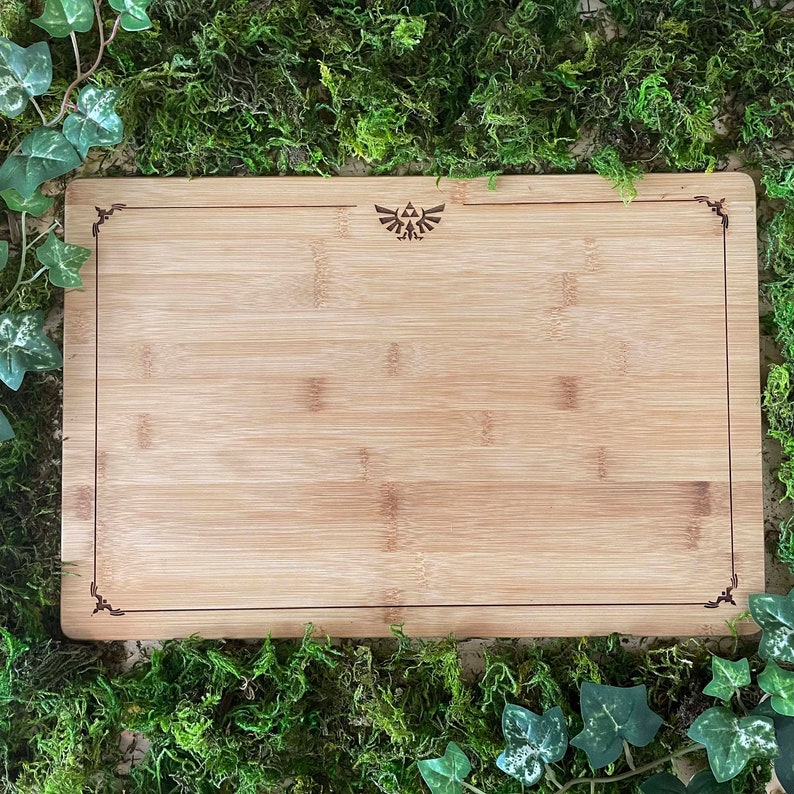 Puede incluir: Una tabla de cortar de bamb&uacute; natural con un borde decorativo y un peque&ntilde;o s&iacute;mbolo triangular grabado en el centro.
