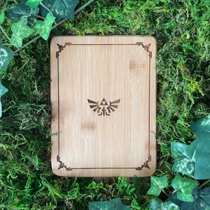 Puede incluir: Una tabla de cortar de madera con un borde decorativo y un s&iacute;mbolo Triforce estilizado en el centro. La tabla est&aacute; hecha de bamb&uacute; marr&oacute;n claro y tiene una veta de madera natural.