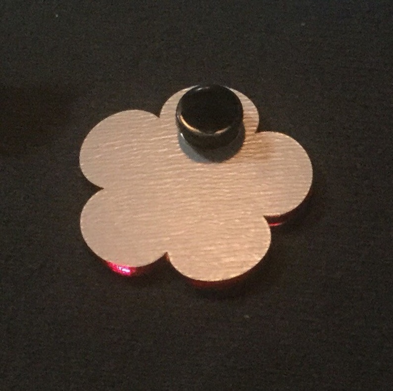 May include: A flower-shaped pin with a light silver finish and a black backing. The pin has a red accent at the bottom and is set against a black background.
