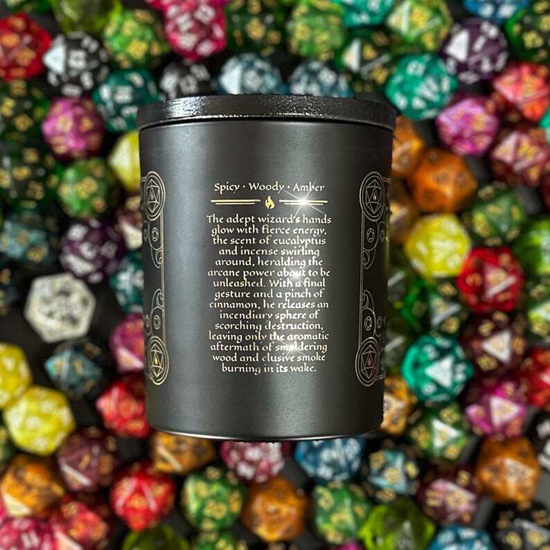 Puede incluir: Un tarro de vela negro con letras doradas. El texto dice "Spicy Woody Amber" y describe las manos de un mago que brillan con una energ&iacute;a feroz, el aroma de eucalipto e incienso que se arremolinan a su alrededor, anunciando el poder arcano a punto de desatarse. Con un gesto final y una pizca de canela, libera una esfera incendiaria de destrucci&oacute;n abrasadora, dejando solo las secuelas arom&aacute;ticas de la madera humeante y el humo esquivo que arde a su paso.