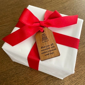 Puede incluir: Una caja de regalo blanca con una cinta roja y una etiqueta de regalo de madera. La etiqueta tiene un dibujo de una pila de regalos y el texto "Make your gift memorable with custom engraved wood tags."