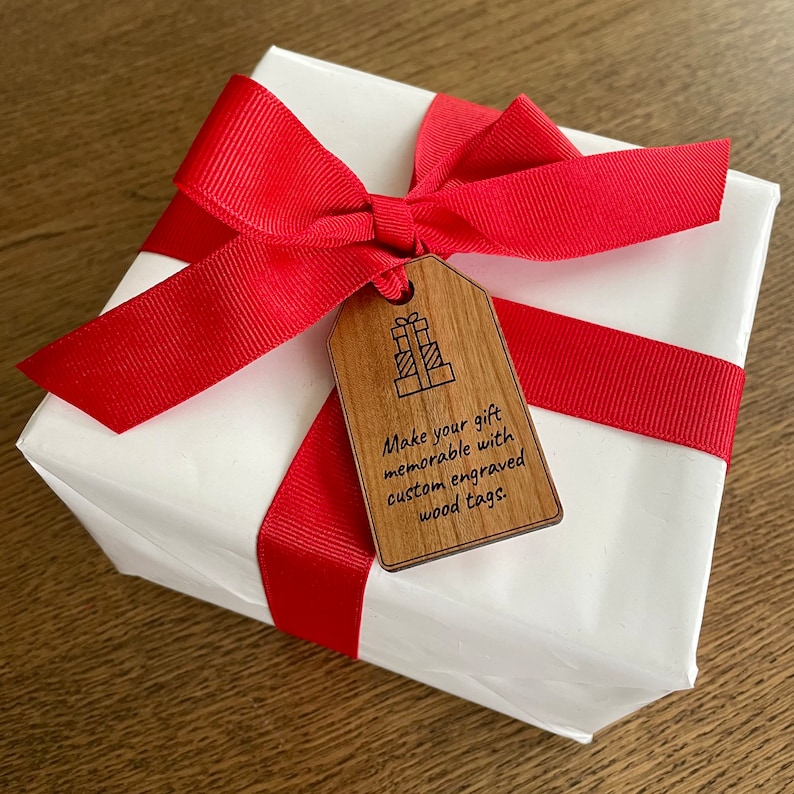 Puede incluir: Una caja de regalo blanca con una cinta roja y una etiqueta de regalo de madera. La etiqueta tiene un dibujo de una pila de regalos y el texto "Make your gift memorable with custom engraved wood tags."