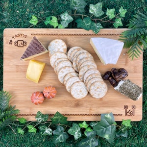 May include: A wooden cutting board with a "So Tasty!" inscription, featuring a variety of cheeses, crackers, and dried fruit. The board has a decorative border and a small engraved design.