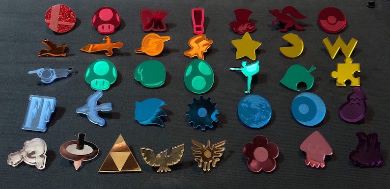 May include: A collection of colorful acrylic pins featuring various shapes and designs. The pins include a mushroom, a star, a Pac-Man, and other video game and pop culture icons. The pins are arranged on a dark gray surface.