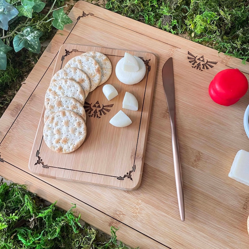 Puede incluir: Una tabla de cortar de madera con un dise&ntilde;o de Triforce, que presenta una variedad de galletas y queso. La tabla es de bamb&uacute; y tiene un acabado natural.
