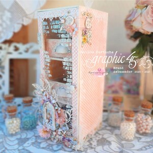 Puede incluir: Una caja decorativa rosa y blanca con un frente de vidrio y un diseño floral. La caja tiene un cajón con una manija de metal. La caja está decorada con encaje, flores y otros adornos. La caja está sentada en una mesa con un mantel blanco. El texto "Graphic 45" es visible en la caja. El texto "Brand Ambassador 2020 - 2021" también es visible en la caja.