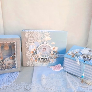 May include: Three blue and white decorated boxes with the name "JUAN BOSCO" on them. The boxes are decorated with lace, flowers, and other embellishments. The largest box is a scrapbook with the text "SCRAPBOOKING CECILIA" on the bottom.