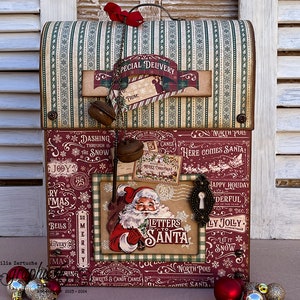 May include: A red and green mailbox with a Santa Claus theme. The mailbox has a sign that says "Letters to Santa" and is decorated with bells, ribbons, and other festive details. The mailbox is sitting on a white wooden surface.