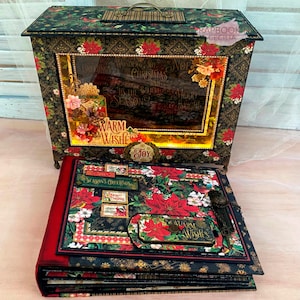 Può includere: Un album di scrapbooking nero e oro con una copertina in velluto rosso e un design floreale rosso e verde. L'album è chiuso e ha un buco della serratura decorativo. L'album è appoggiato su un tavolo di legno. Il testo "Warm Wishes" è visibile sulla copertina.