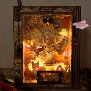 May include: A lighted shadow box with a deer, squirrel, and bird inside. The box is decorated with fall-themed paper and embellishments. The text "Autumn Greetings" is visible on the top of the box. The text "Hello Autumn!" is visible on the bottom of the box.