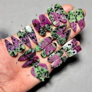 May include: A collection of eleven polished zoisite gemstones, each with a unique pattern. The stones display a mix of colors, including green, pink, and black, with varying shapes and sizes. The gemstones are held in a hand.