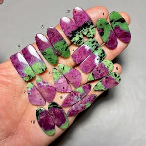 May include: A collection of ten polished ruby zoisite gemstones in various shapes. The stones display a mix of deep pink, green, and black colors. The gemstones are held in a hand, showcasing their natural patterns and textures.