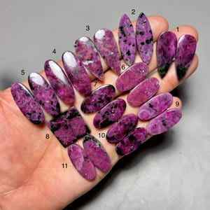 May include: A collection of polished ruby zoisite gemstones in various shapes, including oval, teardrop, and rectangular. The stones display a vibrant pink hue with black and green inclusions, showcasing natural patterns. The gemstones are held in a hand.