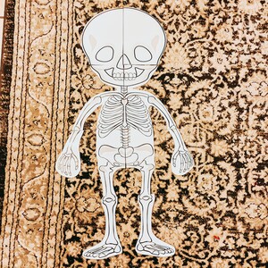 May include: A white paper cutout of a skeleton with a skull head. The skeleton is standing on a brown patterned surface.