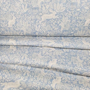 May include: Light blue fabric with a repeating pattern of white rabbits, foliage, and flowers. The fabric is folded, showing multiple layers of the detailed design. The overall theme is a whimsical, nature-inspired design.
