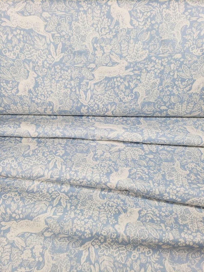 May include: Light blue fabric with a repeating pattern of white rabbits, foliage, and flowers. The fabric is folded, showing multiple layers of the detailed design. The overall theme is a whimsical, nature-inspired design.